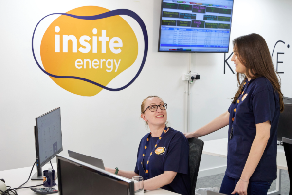 Two Insite Energy customer service team members collaborating at a workstation in the company's contact centre, with computer monitors and operational dashboards visible in the background.