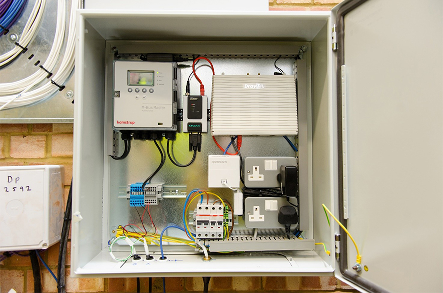 Open electrical control cabinet containing a Kamstrup M-Bus Master unit, a DrayTek router, a Moxa network device, an Openreach termination box, dual power sockets, circuit breakers, and neatly organised wiring.