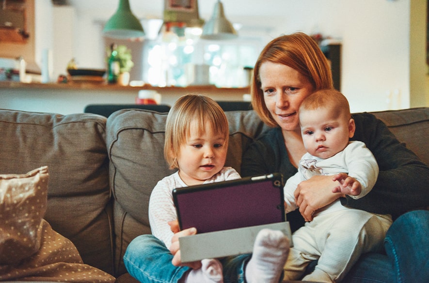 Adult seated on a sofa indoors with two young children, holding a tablet while the children look at the screen, in a living room setting with cushions and kitchen area visible in the background.
