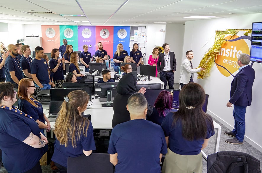 Group of people gathered in an open‑plan office around desks and computer workstations, watching two people unveil an Insite Energy wall sign by removing gold material. Insite Energy branding is visible on the wall, with service team signage displayed along the back of the room.