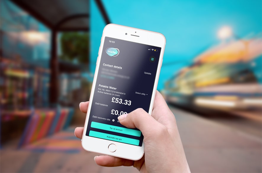 Smartphone held in one hand displaying the my insite energy app account screen, showing contact details, a potable water balance of £53.33, and buttons labelled “Top up account” and “Automatic top‑ups,” with a blurred street and bus stop visible in the background.