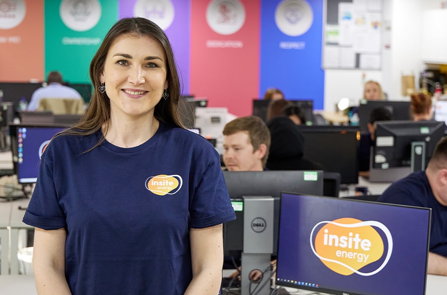 Person wearing an Insite Energy T‑shirt standing in an open‑plan office, with desks, computer monitors displaying the Insite Energy logo, and other people working at workstations in the background.