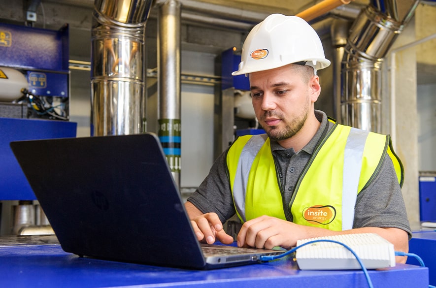 Person wearing a hard hat and high‑visibility vest using a laptop on a work surface inside a plant room, with pipes, insulated equipment, and Insite Energy branding visible.