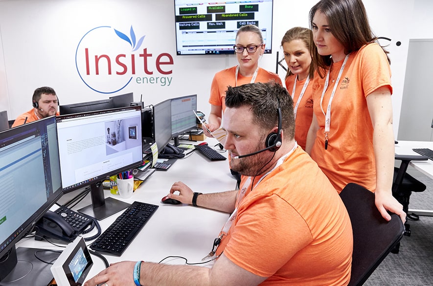 People working at computer desks in a contact centre environment, with one person seated wearing a headset and using multiple monitors while others stand nearby observing the screen. Insite Energy branding is visible on the wall behind the desks, with call‑handling software displayed on monitors and office equipment arranged across the workspace.