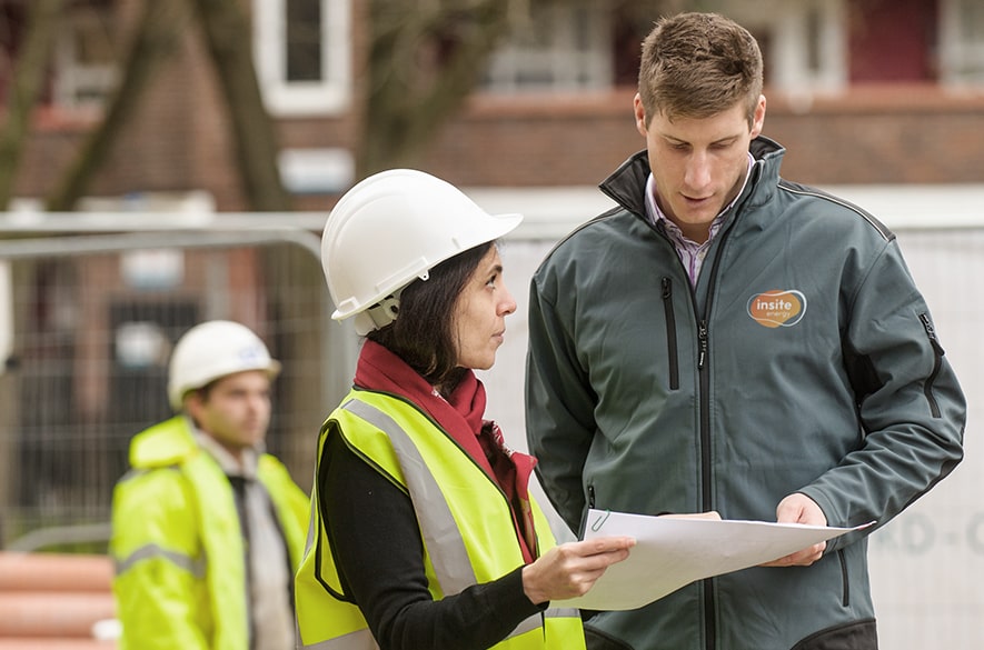 Two people wearing safety helmets and high‑visibility clothing reviewing a document together at an outdoor construction site, with fencing and another person in protective clothing visible in the background.