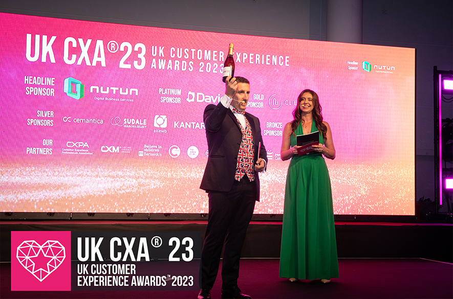 Stage scene at the UK CXA 2023 event, showing two presenters standing under a large screen displaying “UK CXA® 23 UK Customer Experience Awards 2023” with sponsor logos, while one presenter holds an award above their head.