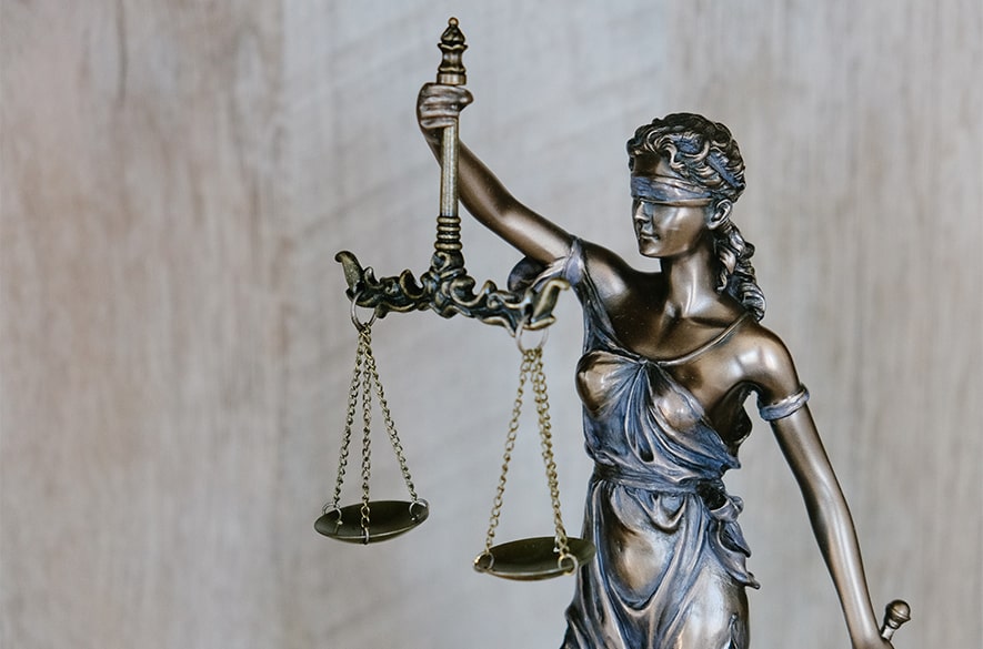 Close‑up of a bronze statue of Lady Justice holding a set of balanced weighing scales in one hand. The figure wears a blindfold and stands against a plain stone background, with detailed metalwork visible on the scales, chain, and clothing.
