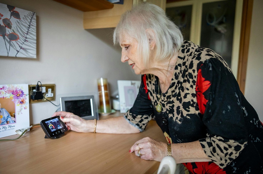 Person leaning over a kitchen counter adjusting a small smart heating control device with a digital display, placed on a wooden worktop. Framed photographs, a tablet device, and household items are visible on the counter and shelves nearby.