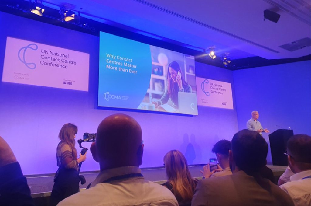Conference presentation at the UK National Contact Centre Conference, showing a speaker on stage and an audience seated in the foreground. Large presentation screens display the text “Why Contact Centres Matter More than Ever” alongside UK National Contact Centre Conference branding, with stage lighting and cameras visible in the room.