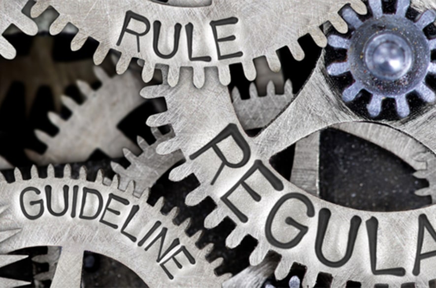 Close‑up of interlocking metal gears, with the words “Rule,” “Regulation,” and “Guideline” engraved on different gear cogs.