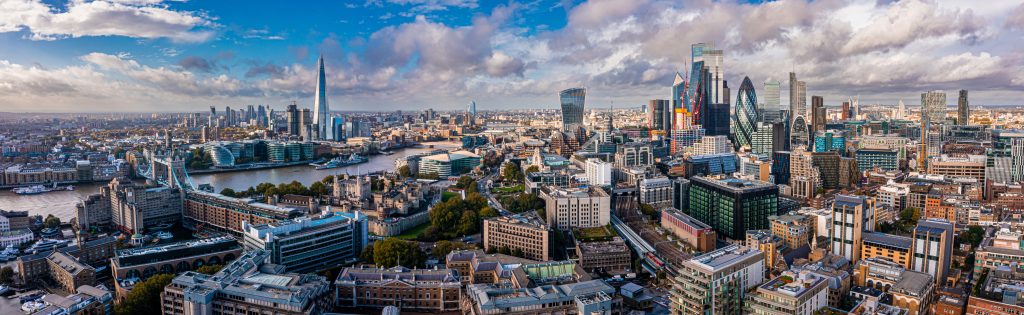 Panoramic aerial view of central London, showing the River Thames, Tower Bridge, and a dense city skyline with modern and historic buildings under a partly cloudy sky.
