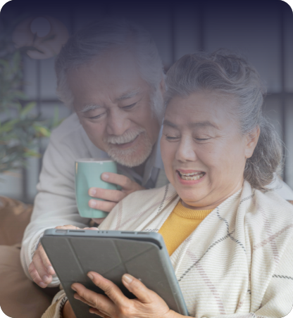 Two people seated close together indoors, one holding a tablet while the other looks at the screen and holds a mug.