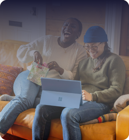 Two people seated on a sofa using a tablet, with one holding a patterned gift bag. Cushions and a laptop-style tablet are visible in a living room setting.