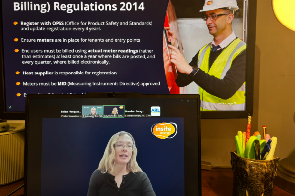A desk setup with two screens: one shows a presentation slide titled “Billing Regulations 2014” alongside an image of a worker in safety gear inspecting equipment, and the other shows a video call with Insite's Commercial Director and the Insite Energy logo.