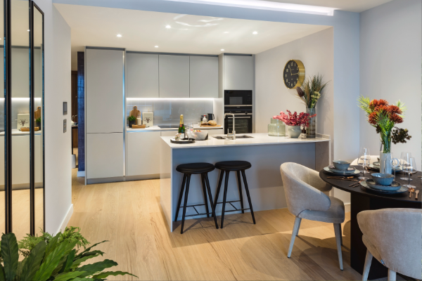 Interior view of an apartment at South Quay Plaza, showing an open‑plan kitchen and dining area with fitted units, an island with bar stools, built‑in appliances, and a dining table with chairs. Decorative plants, table settings, and wall decor are visible, with wood flooring and recessed ceiling lighting throughout.