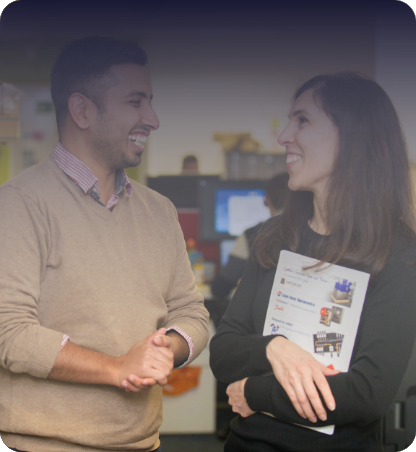 Two colleagues discussing documents while standing beside shelving, with a blurred workspace in the background.